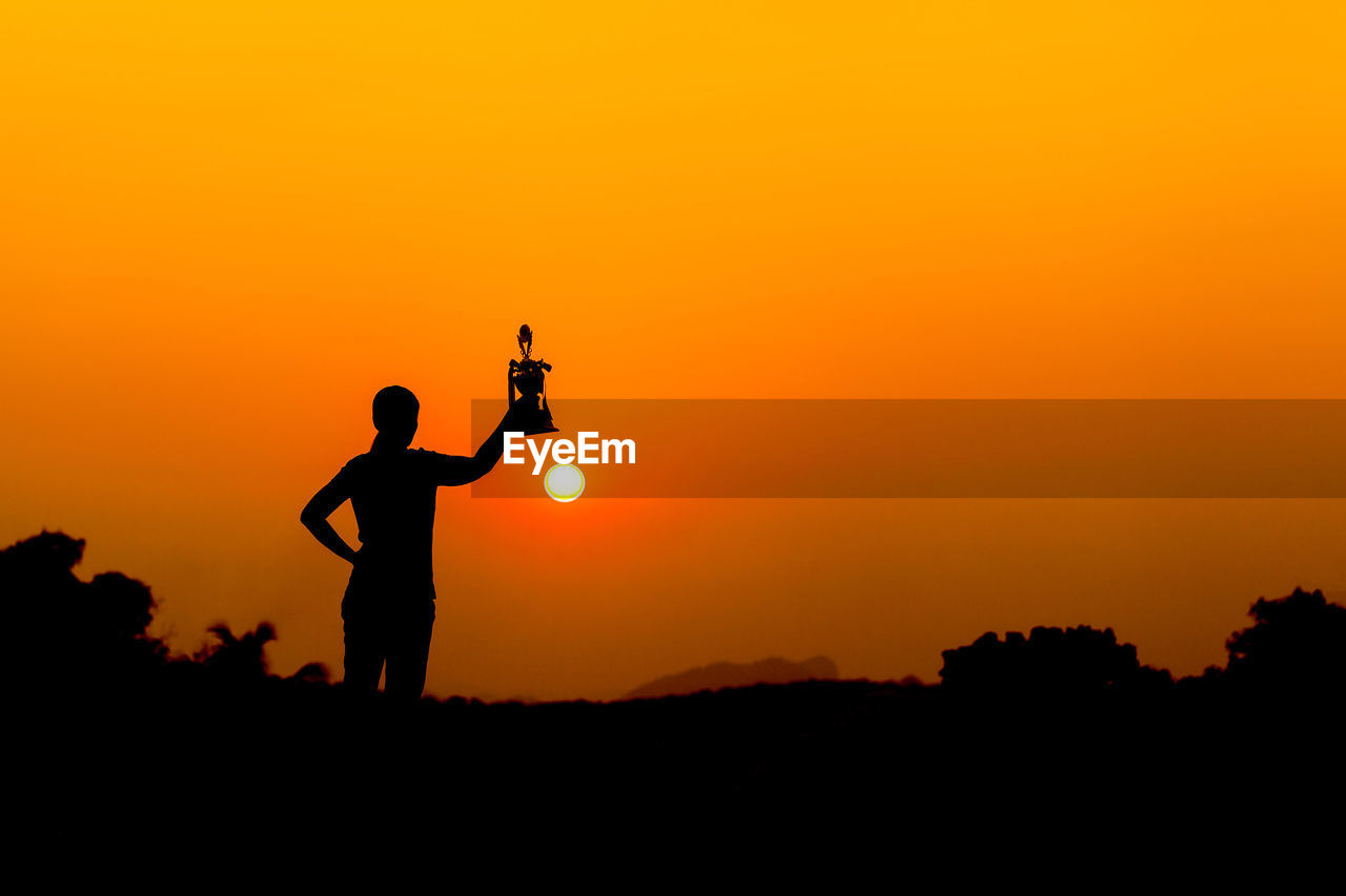 Silhouette woman holding trophy while standing on land against sky during sunset