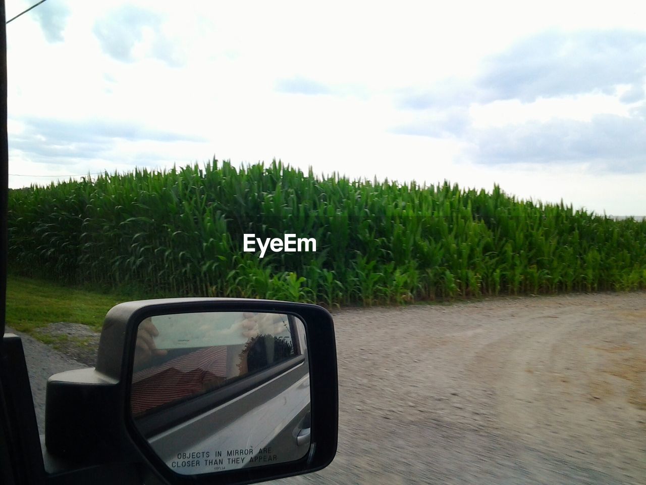 Cropped image of car side-view mirror by agricultural against sky