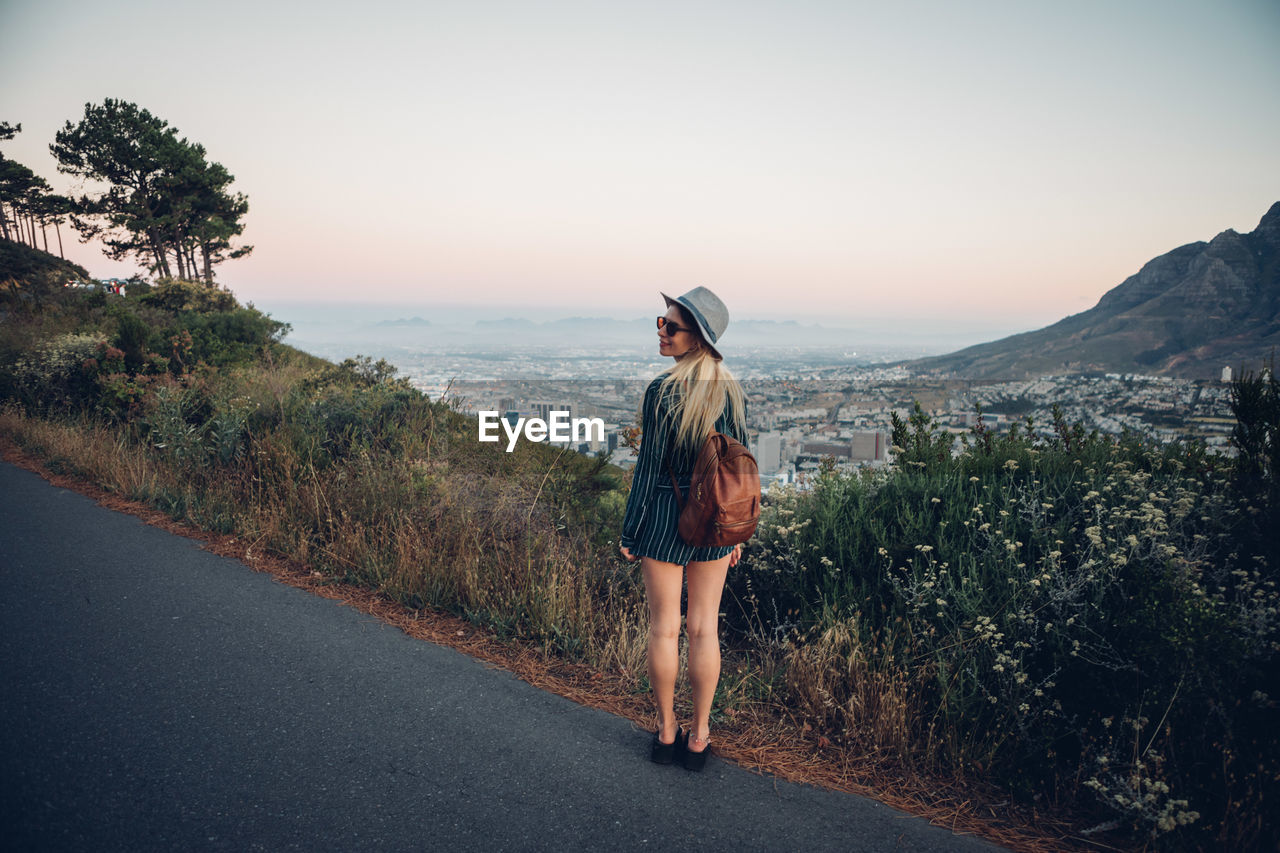 Rear view of woman standing on road by cityscape against clear sky during sunset