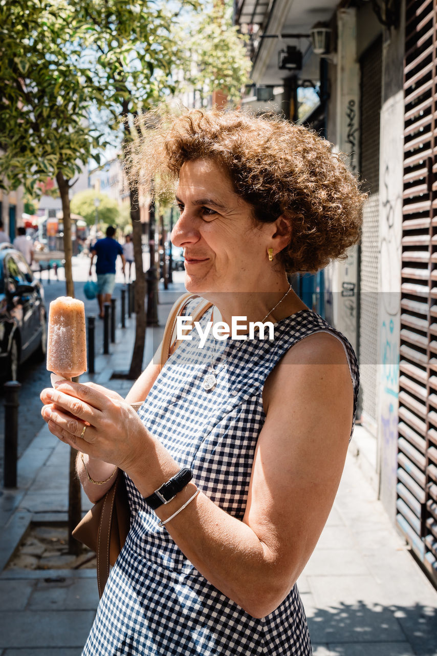 Mature woman with popsicle looking away while standing on sidewalk in city