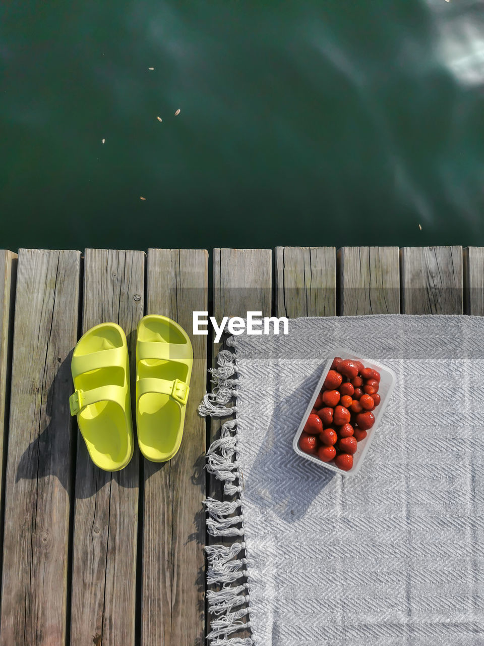 Neon sandals, flip flops with beach rug and box of strawberries on wooden floor beside lake water.