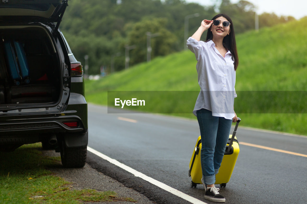 Full length of woman wearing sunglasses with suitcase standing on road