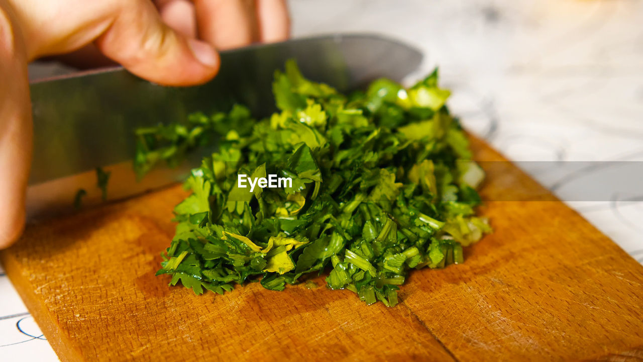 A close-up of a woman's hand with a knife is chopping parsley on a cutting board.