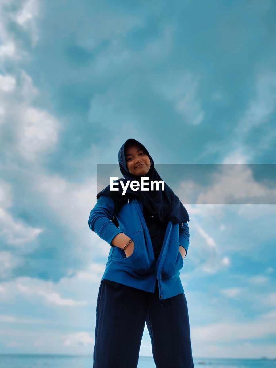 Low angle portrait of woman wearing hijab standing against cloudy sky