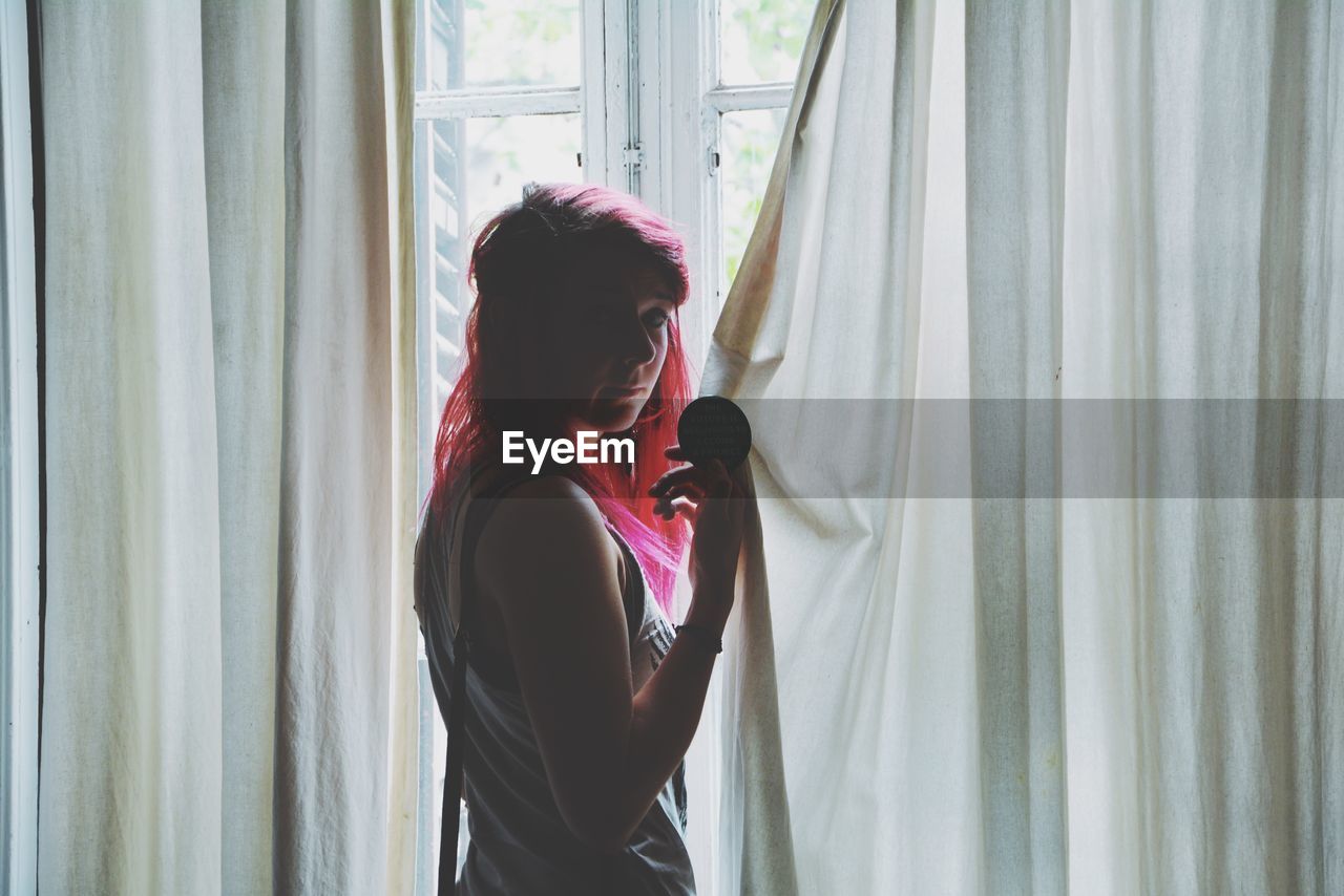 Woman with pink hair standing by curtain against window at home