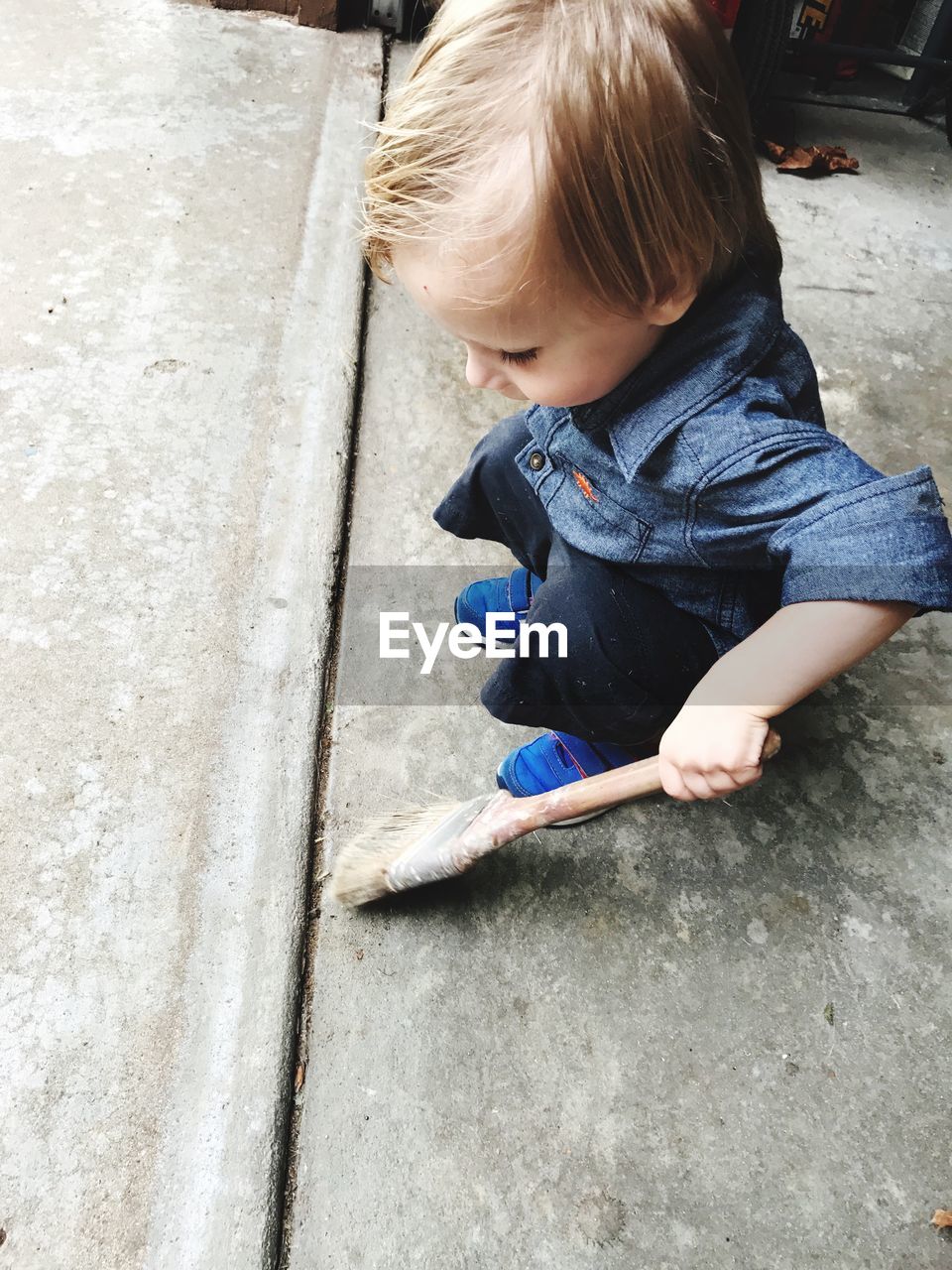 High angle view of boy holding paintbrush while crouching on footpath