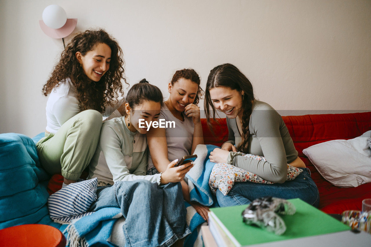 Happy young female friends looking in smart phone while sitting on sofa at home