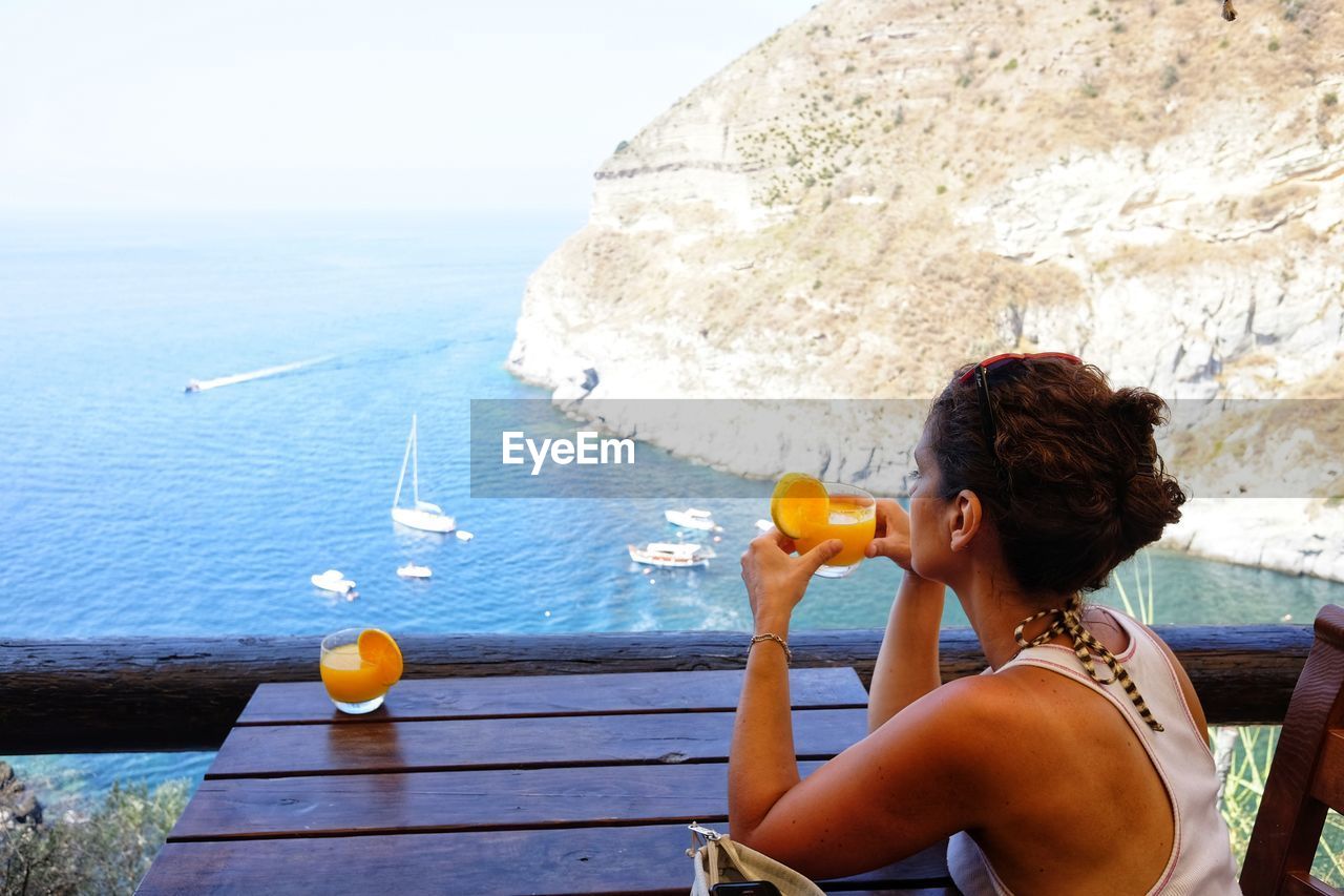 Side view of woman holding orange juice while looking at sea from restaurant