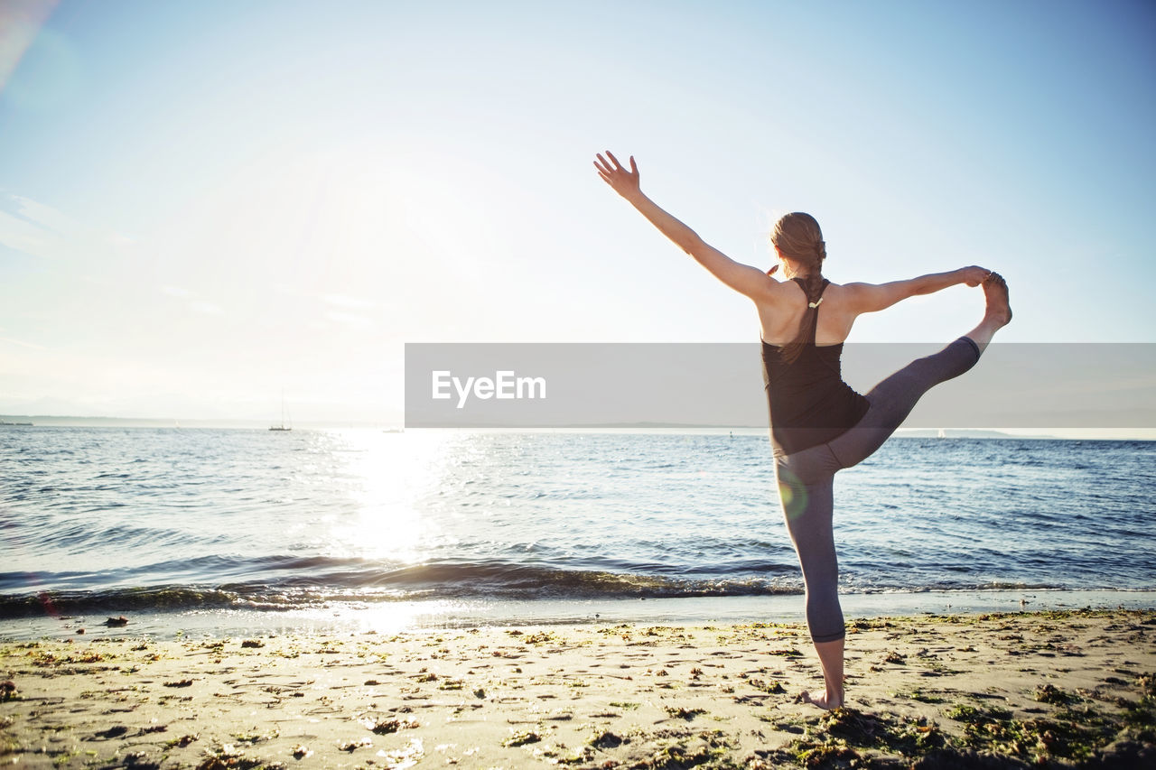 Rear view of woman practicing hand to big toe yoga pose at beach during sunny day