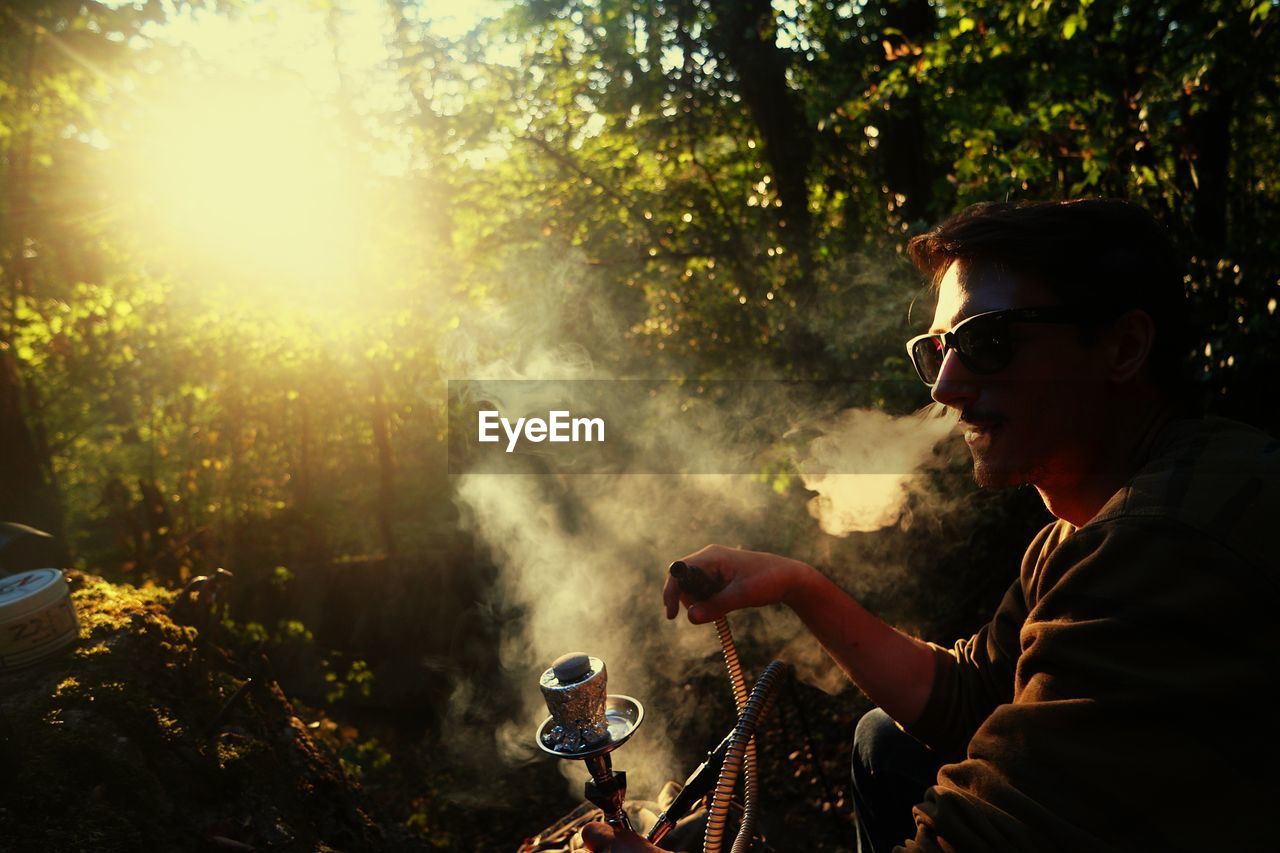 Side view of young man smoking hookah in forest during sunset