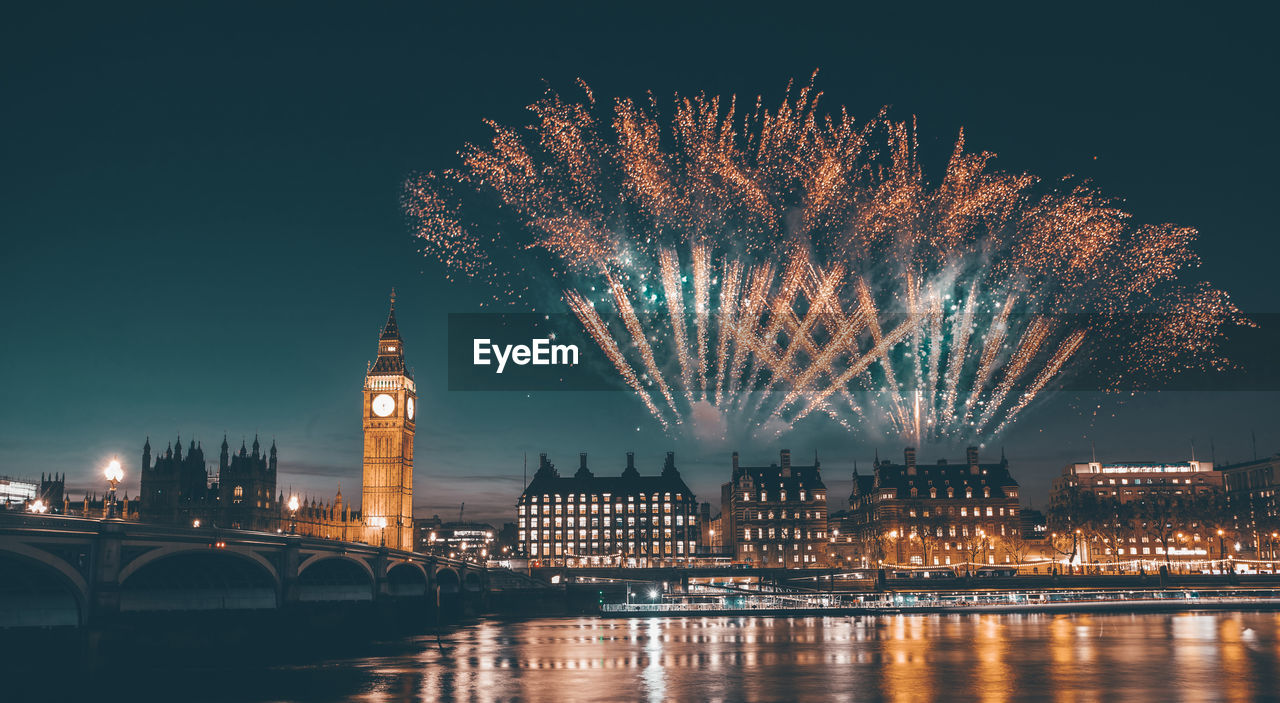 FIREWORK DISPLAY OVER RIVER WITH ILLUMINATED BUILDINGS IN BACKGROUND AT NIGHT