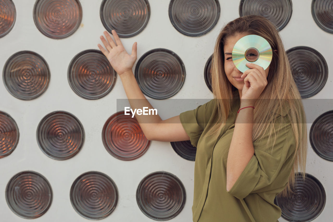 Portrait of woman looking through compact disc while standing against wall