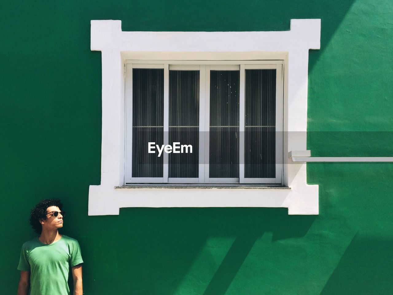 Young man looking away while standing by closed window on green wall