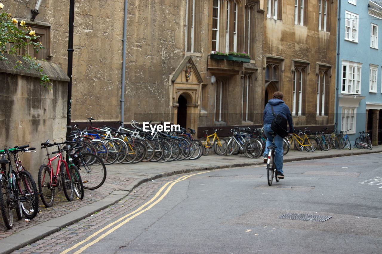 Rear view of man riding bicycle on road with bycicles parked against the buildings.