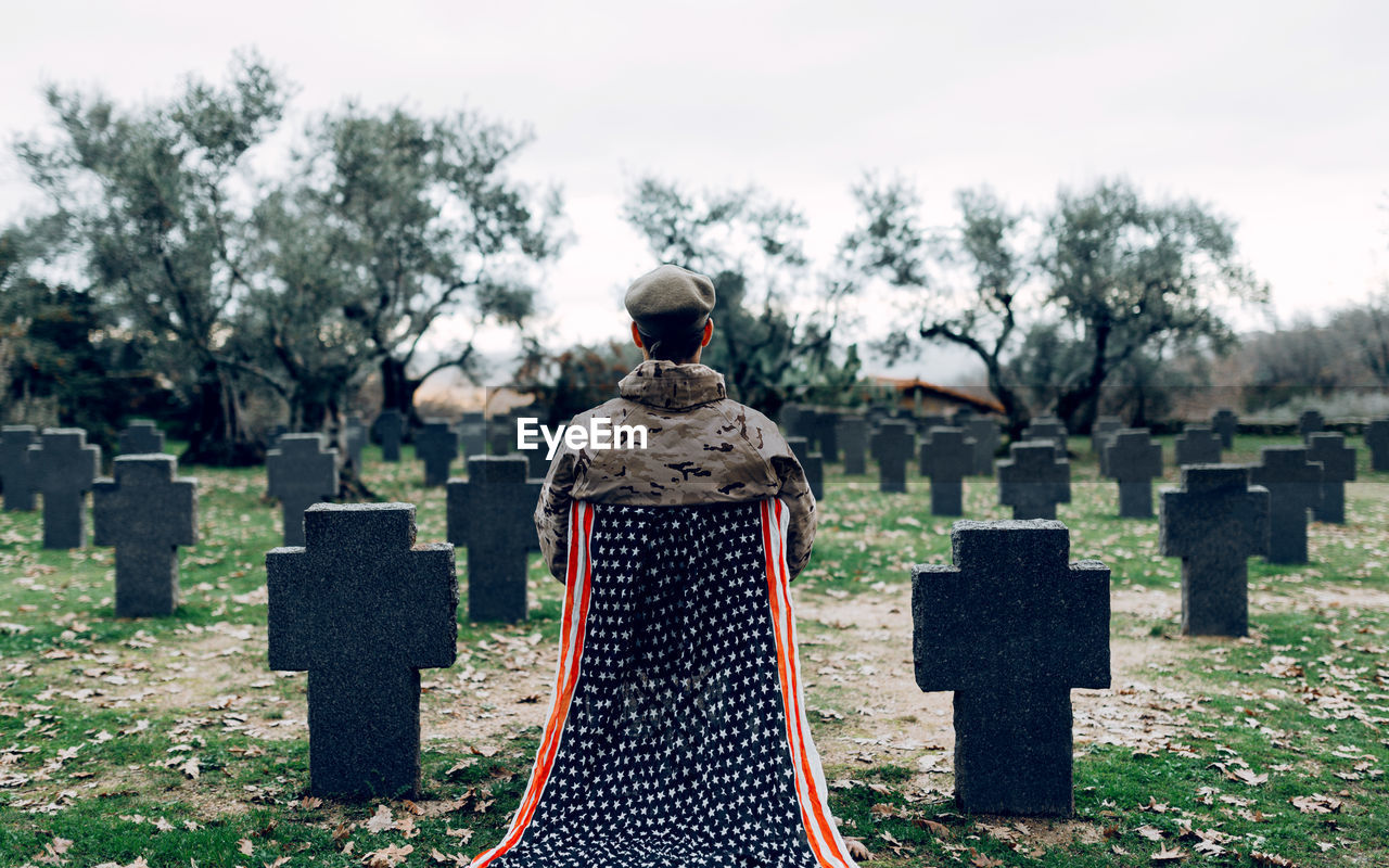 Back view full body of soldier in uniform sitting on chair with american flag while mourning death of warriors at graveyard