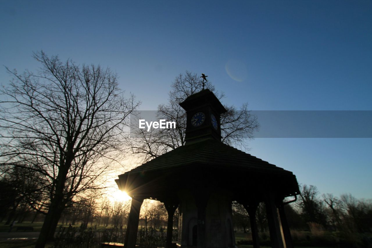 Low angle view of clock tower by silhouette bare trees against clear blue sky during sunset