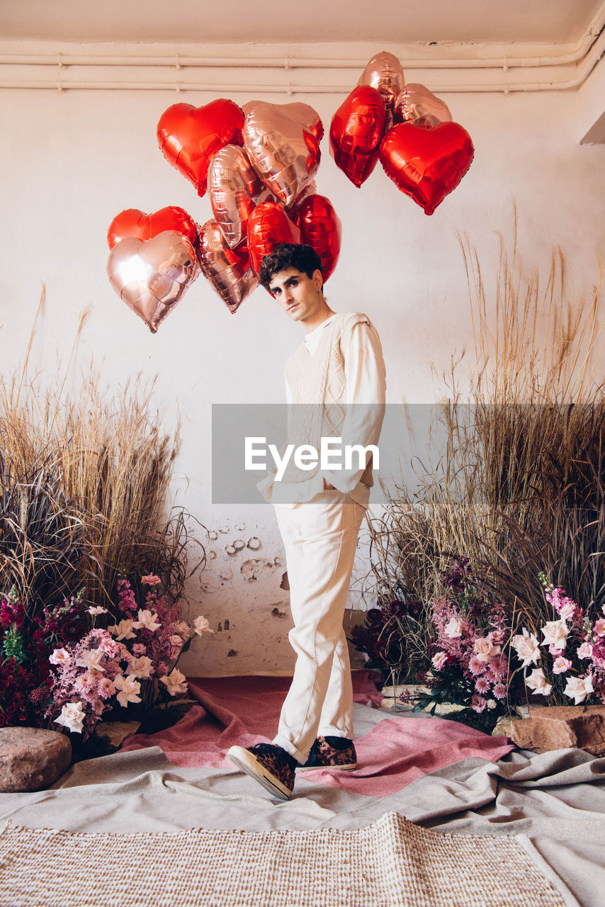 Full body of hispanic male in stylish white wear looking at camera while standing near colorful heart shaped balloons and flowers