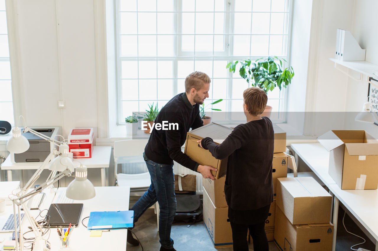 High angle view of business colleagues stacking cardboard boxes by window in creative office