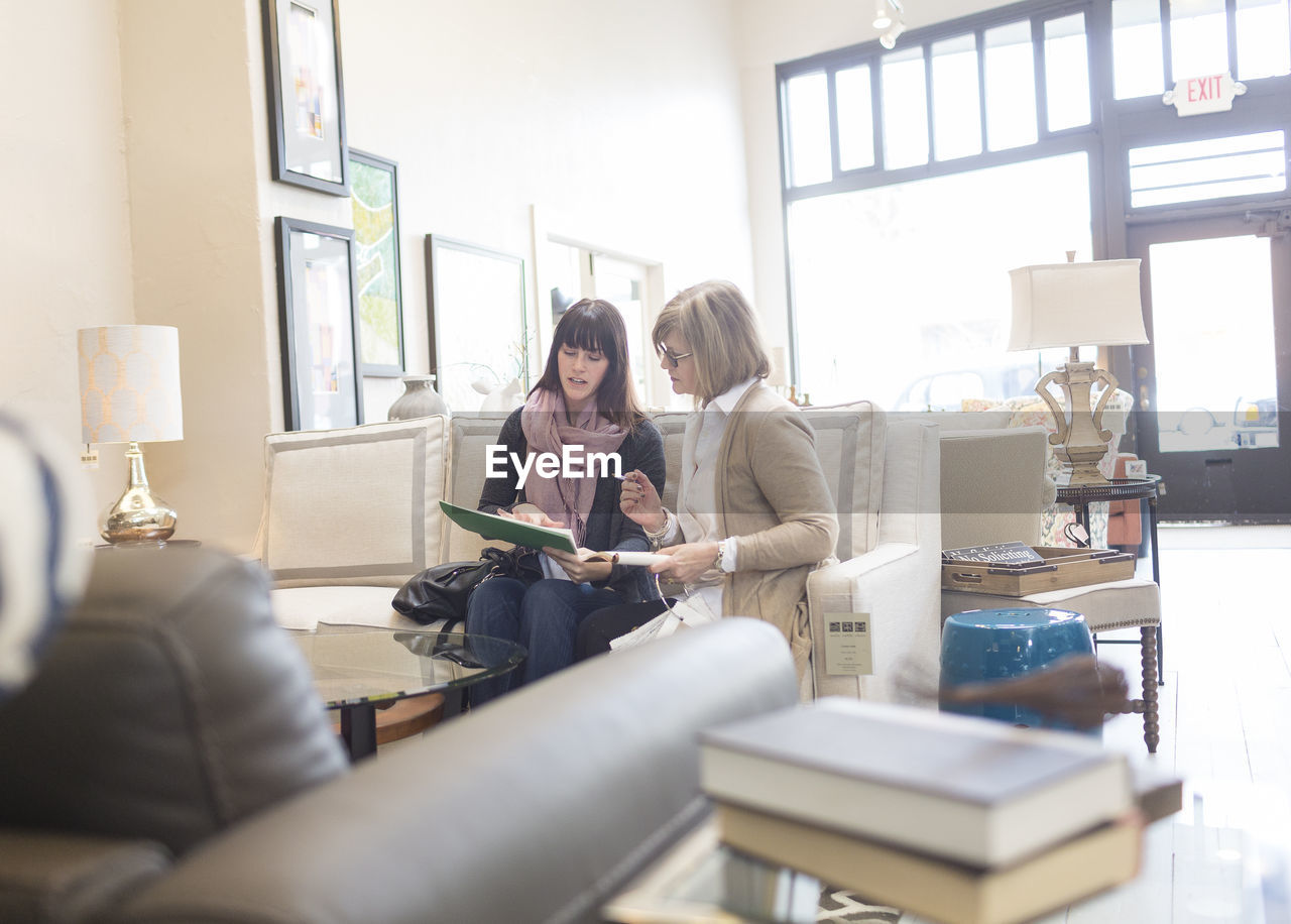 Saleswoman discussing with customer while sitting on sofa in furniture store