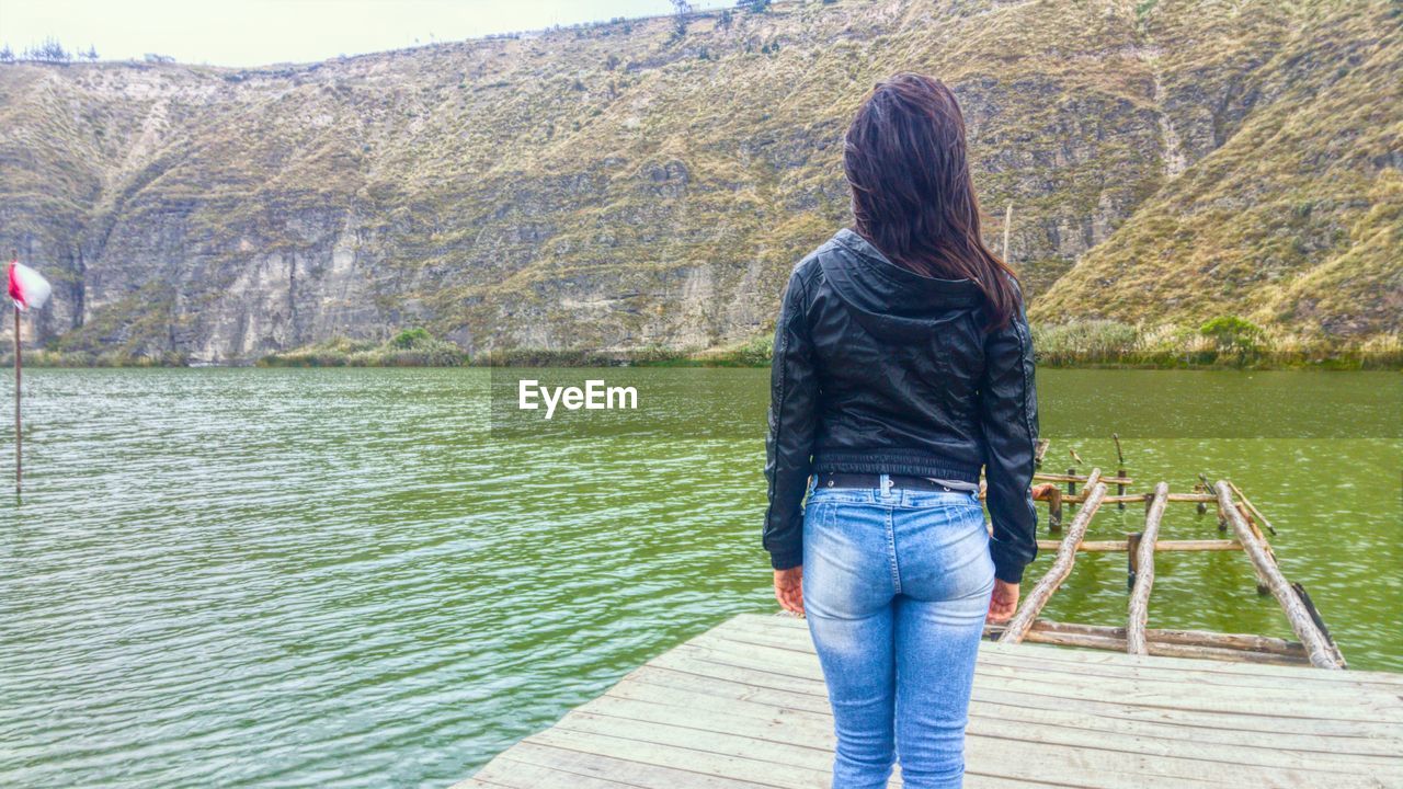 Rear view of young woman standing on pier over lake amidst mountain