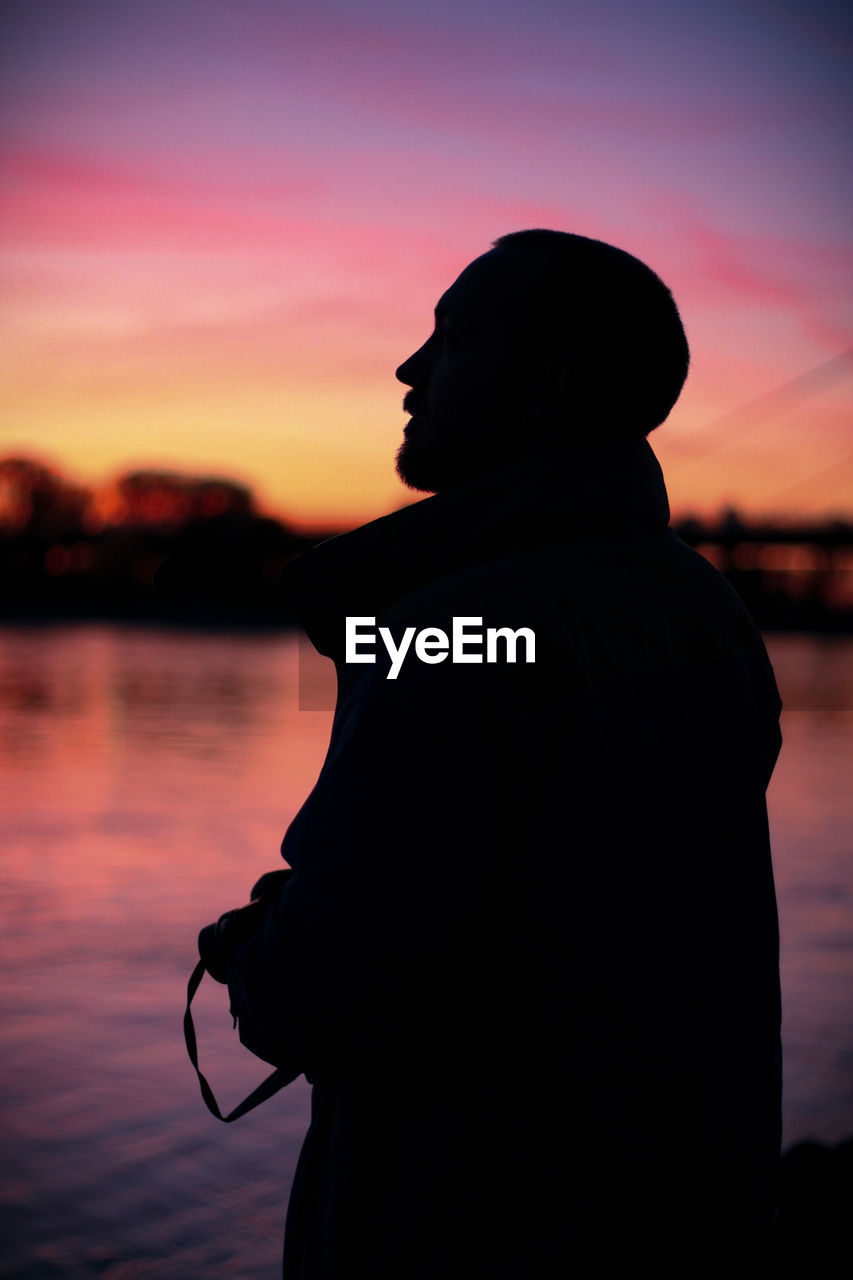 Silhouette of man standing by river against sky during sunset