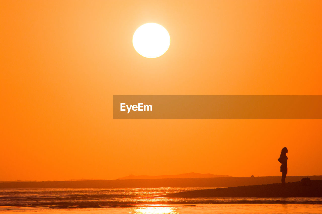 SILHOUETTE WOMAN STANDING AT BEACH AGAINST ORANGE SKY