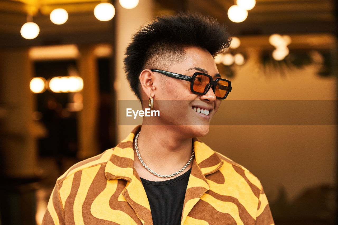 Smiling young non binary male in stylish shirt with sunglasses and accessories with face skin imperfections looking away