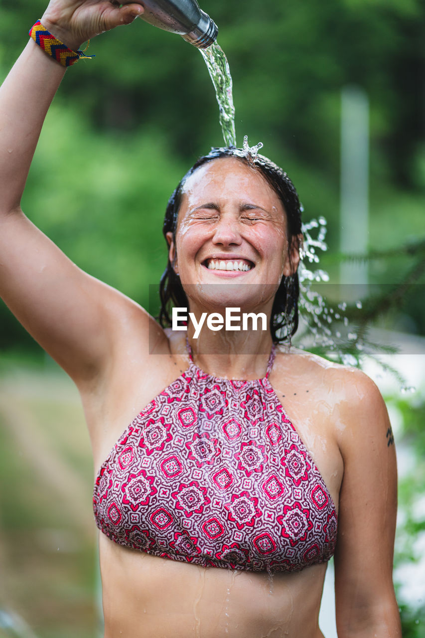 Happy female standing with closed eyes in top and pouring water from bottle on head against green trees in park in summer time