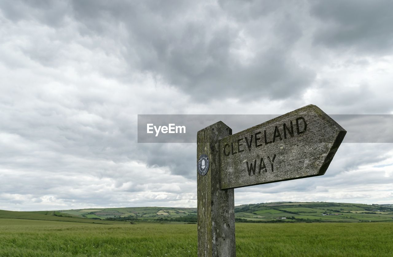 Low angle view of directional sign on green landscape against cloudy sky