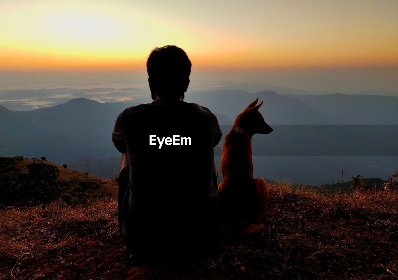 REAR VIEW OF MAN LOOKING AT MOUNTAIN DURING SUNSET