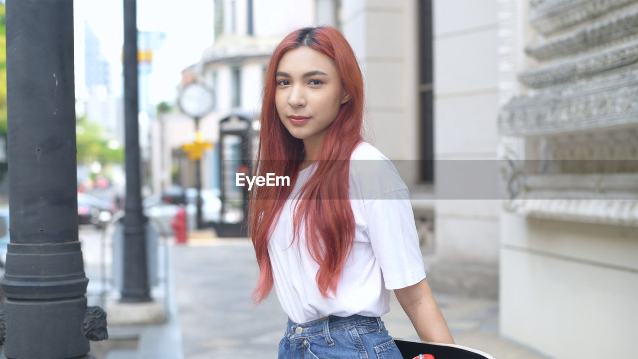 PORTRAIT OF BEAUTIFUL YOUNG WOMAN STANDING AGAINST CITY IN BACKGROUND