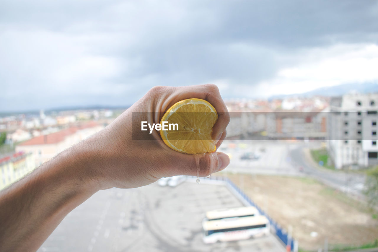 Cropped hand of man squeezing lemon against cityscape