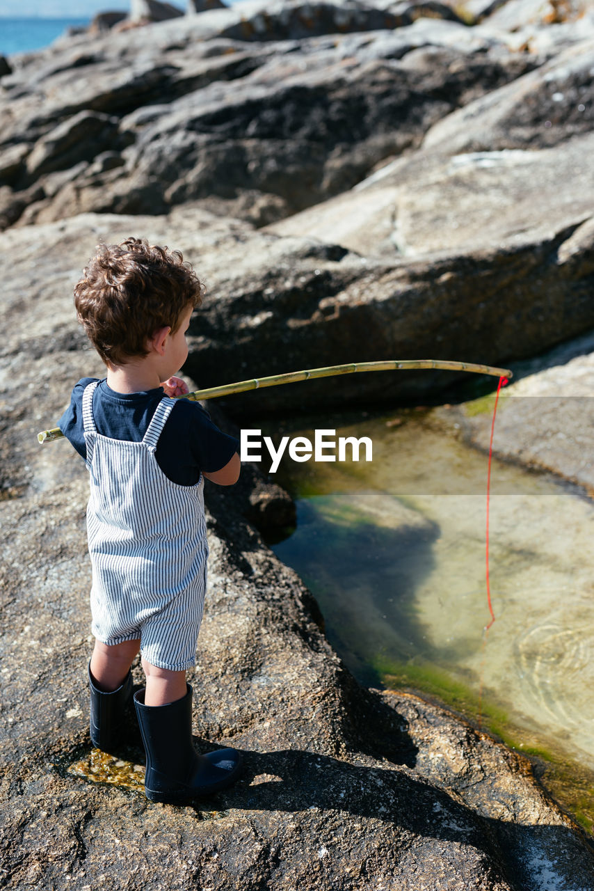 Side view of adorable child fisher in gumboots and overall holding small rod and fishing in shallow clear brook on rocks