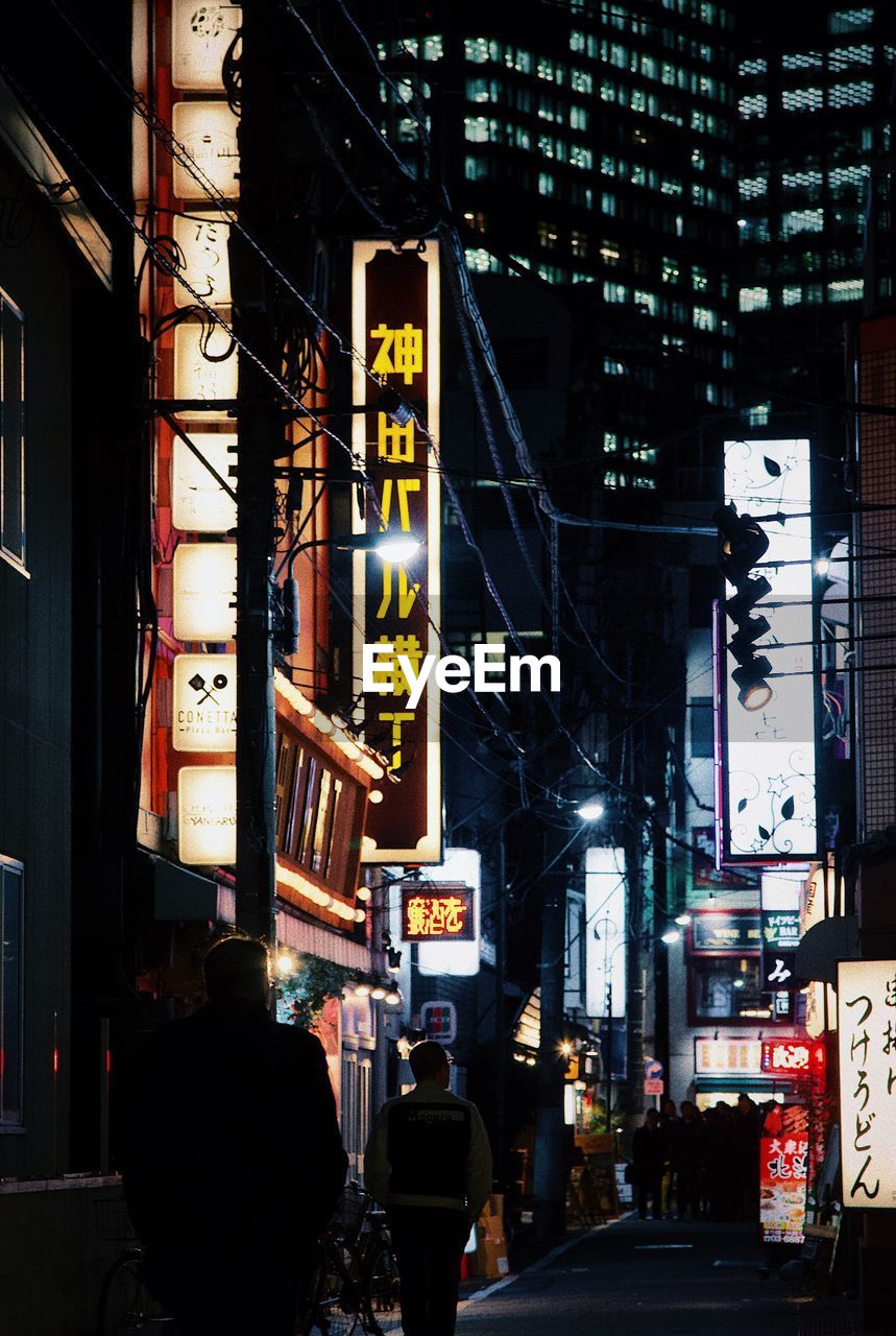 PEOPLE ON ILLUMINATED STREET AMIDST BUILDINGS AT NIGHT