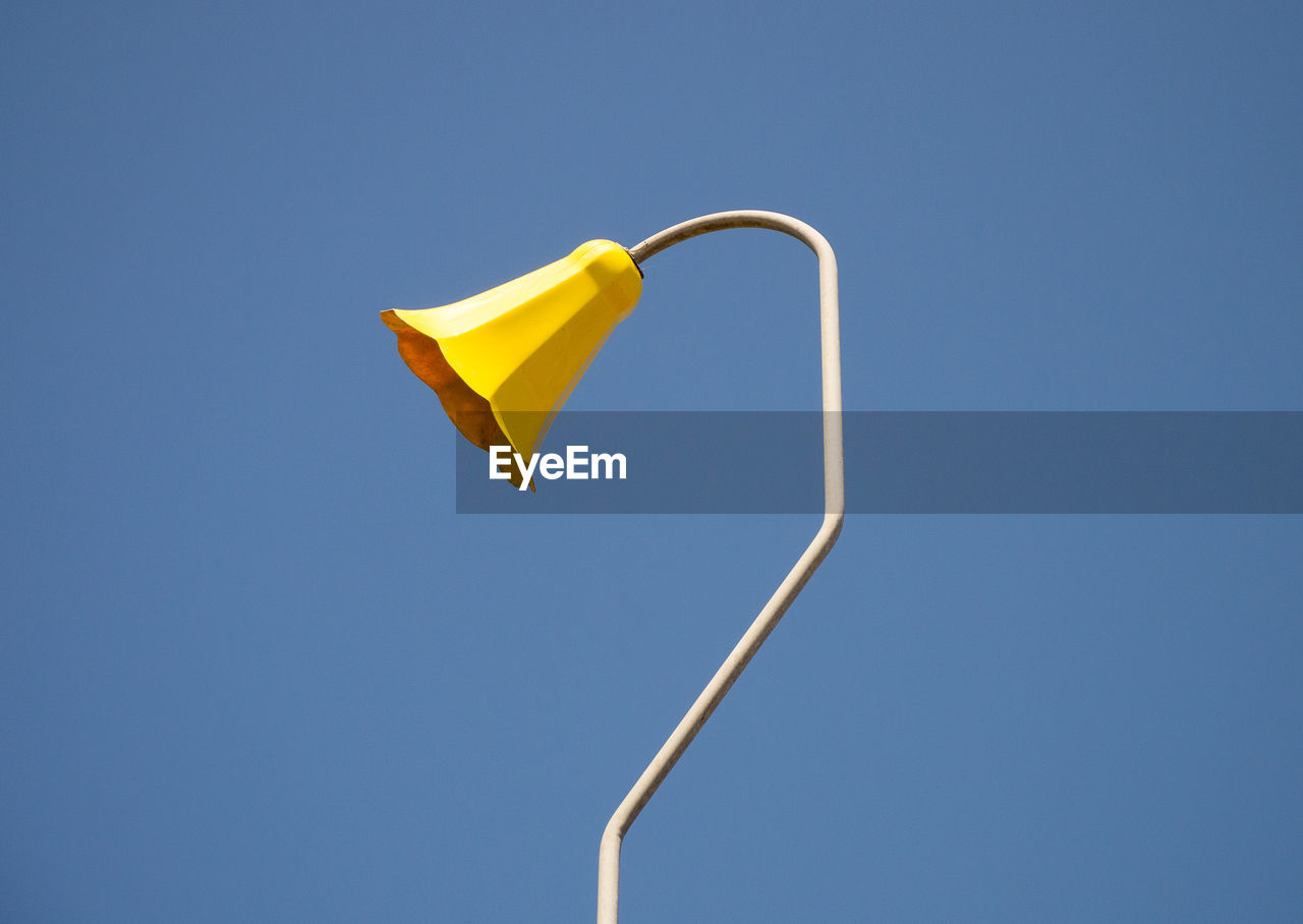 An old fashioned yellow flower-shaped lamp on a curved lamp post, against a clear blue sky
