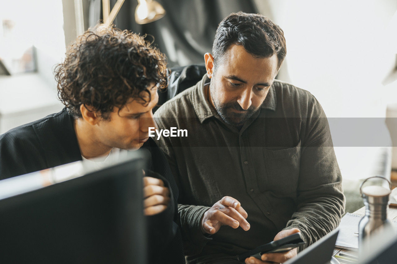 Male it professional explaining ideas to colleague while using smart phone in tech startup office