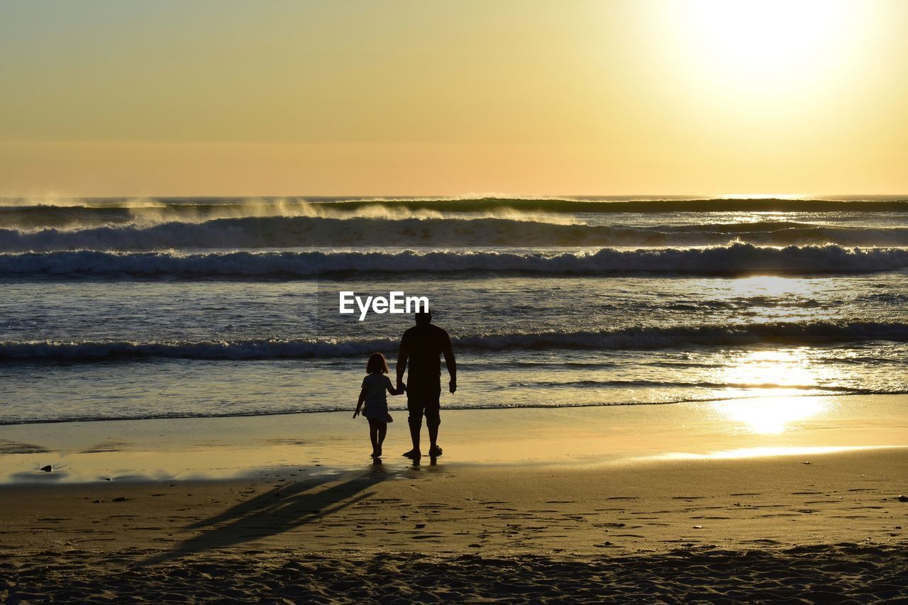 silhouette couple walking at beach against sky during sunset