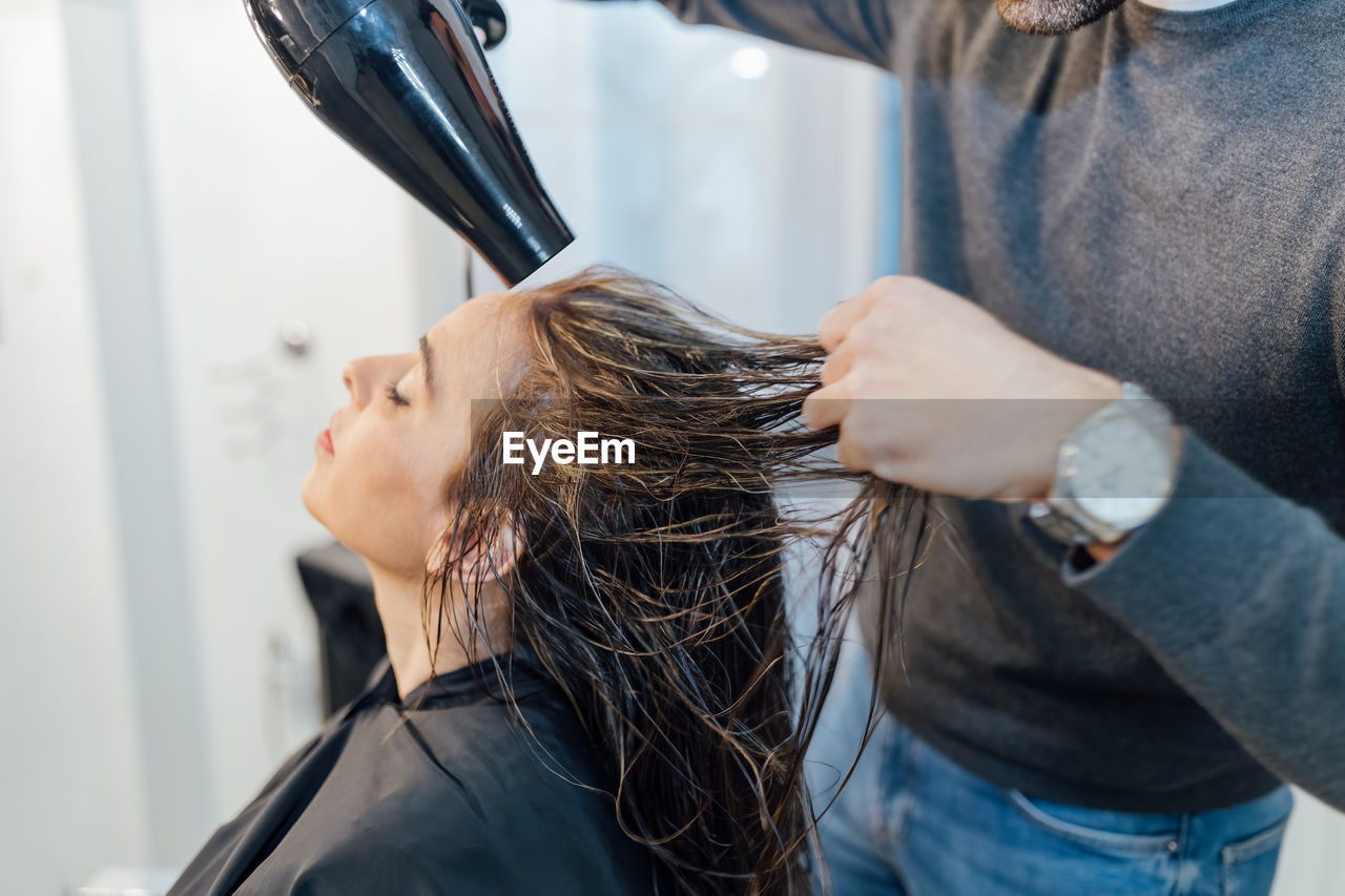 Crop young ethnic male hairdresser drying hair of female client with closed eyes in modern beauty studio