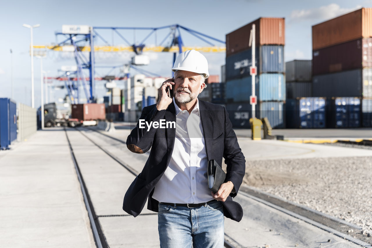 Businessman at cargo harbour, wearing safety helmet, using smartphone