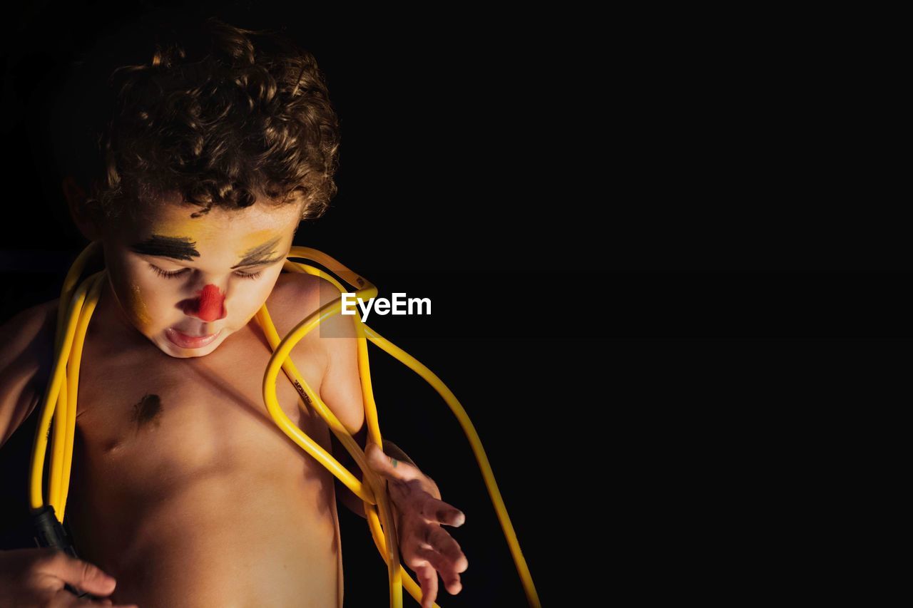 Shirtless boy with face paint and yellow cables standing against black background