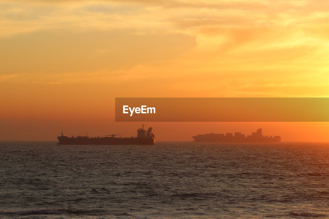 Silhouette container ships in sea against orange sky during sunset