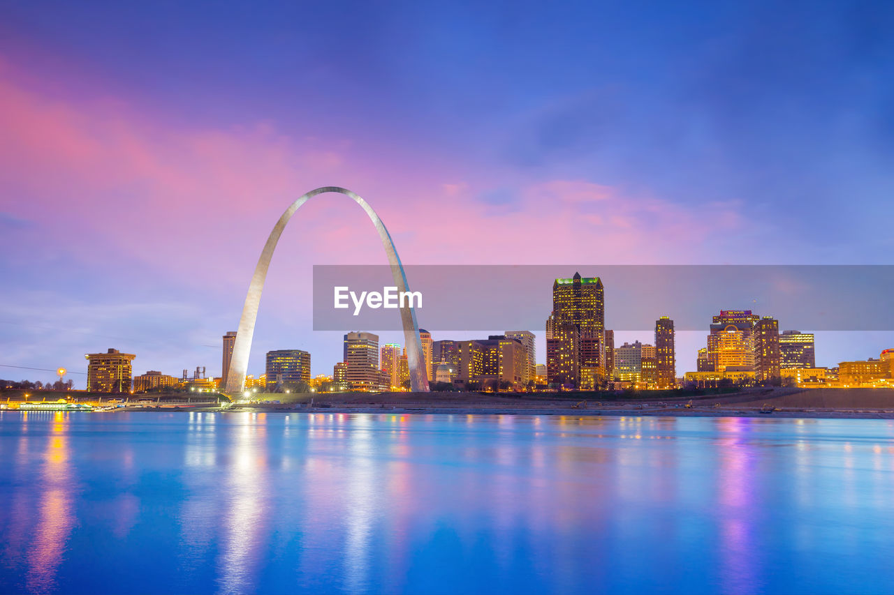 ILLUMINATED BUILDINGS BY RIVER AGAINST SKY IN CITY DURING SUNSET