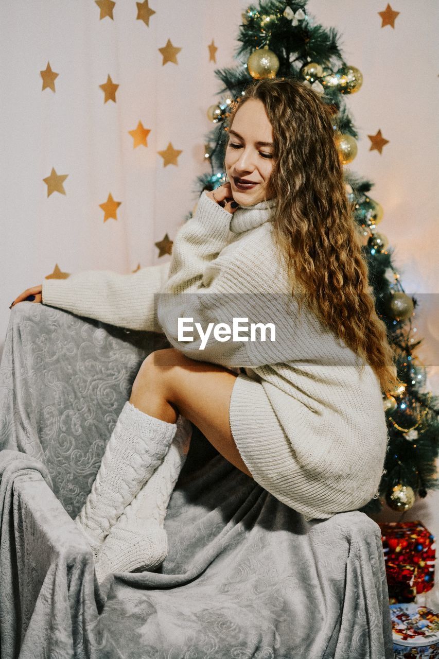 Smiling woman sitting on chair against christmas tree at home