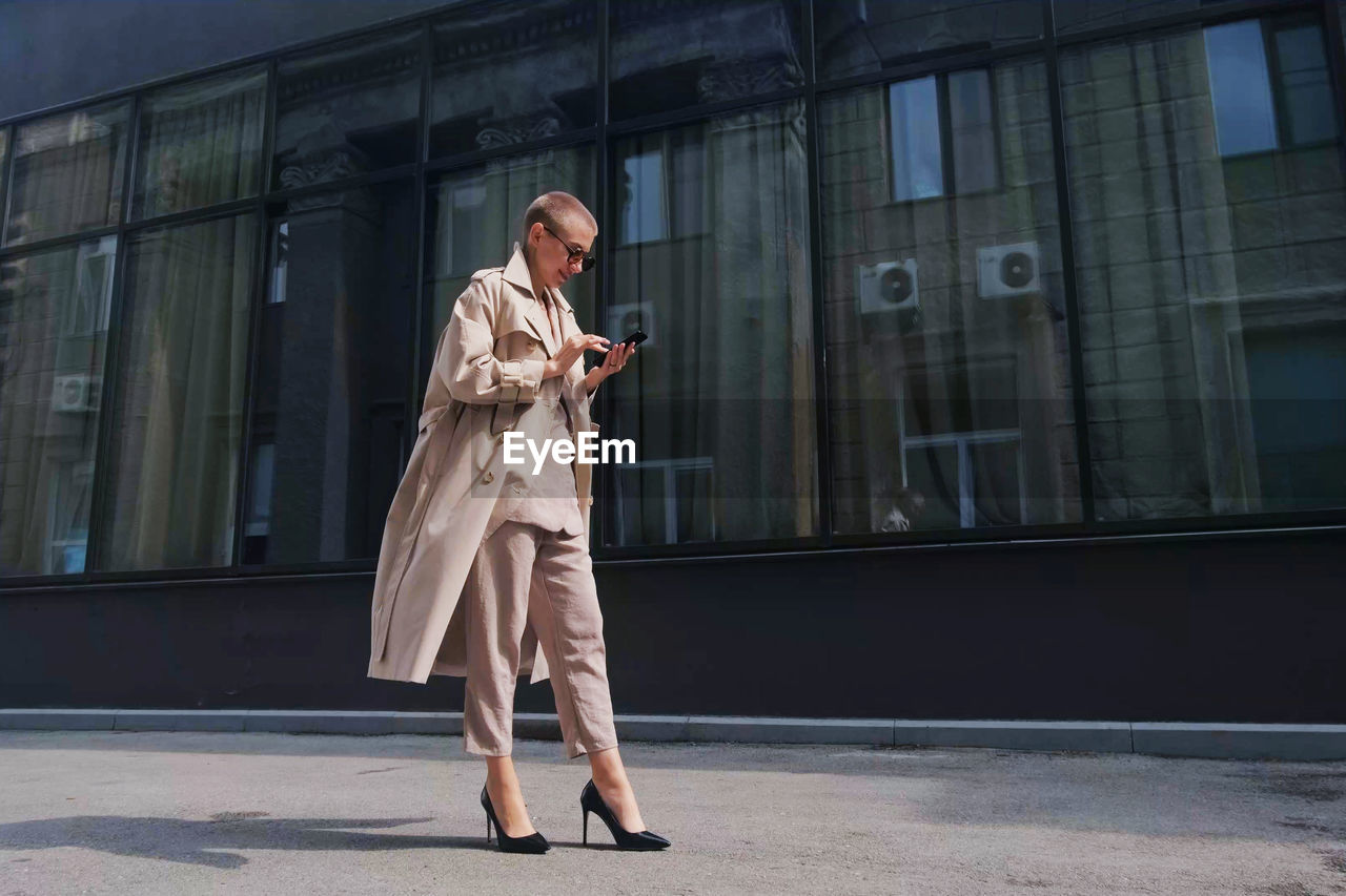 Full length of businesswoman using mobile while standing against building