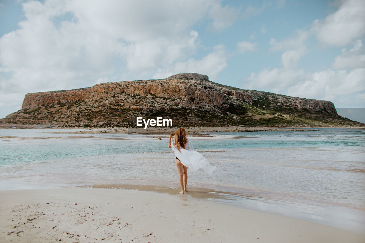 Full length slim female in black swimsuit standing on sandy balos beach against rocky cliff and looking away on clear sunny weather