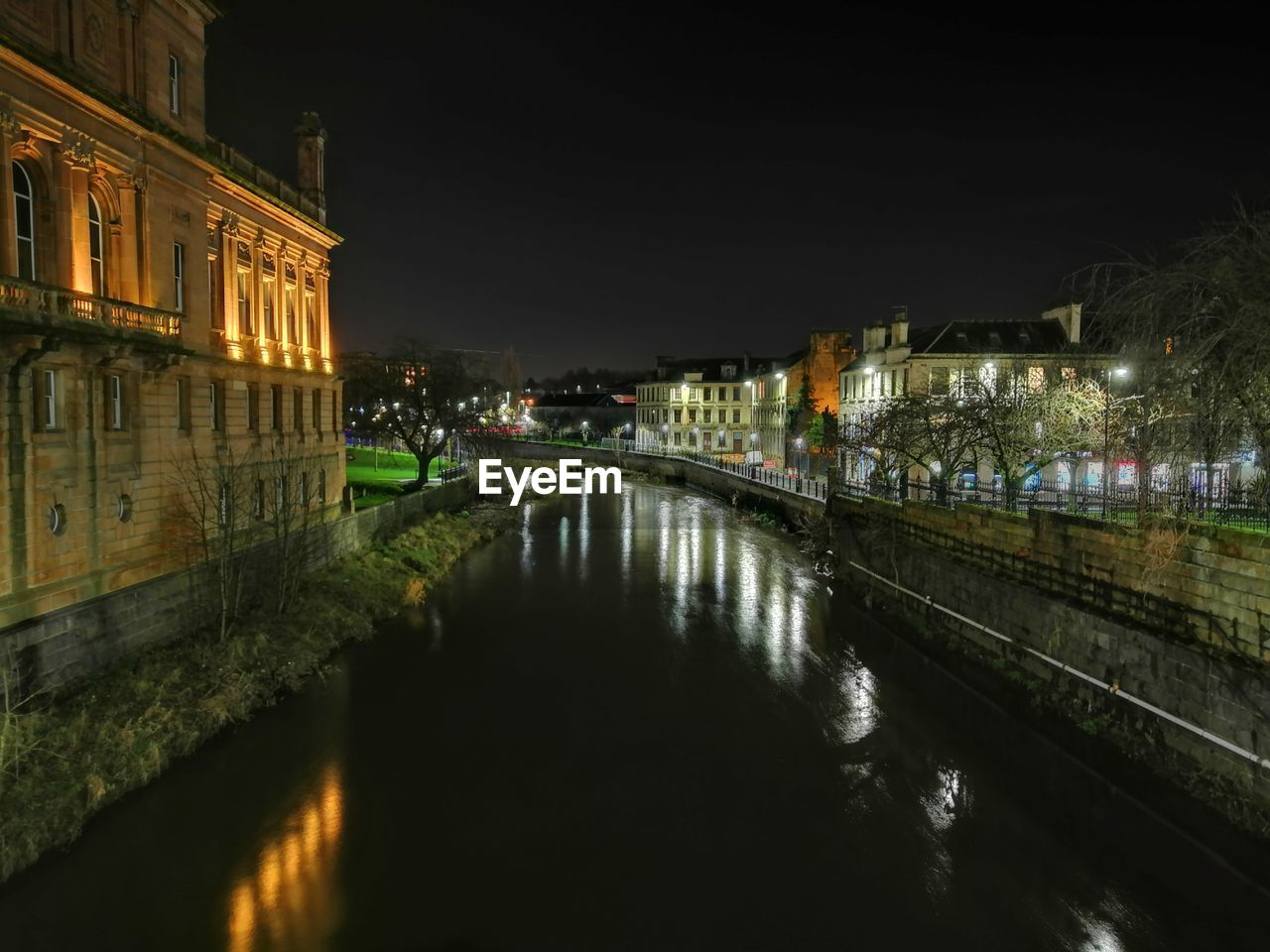 ILLUMINATED BRIDGE OVER RIVER BY BUILDINGS AGAINST SKY AT NIGHT