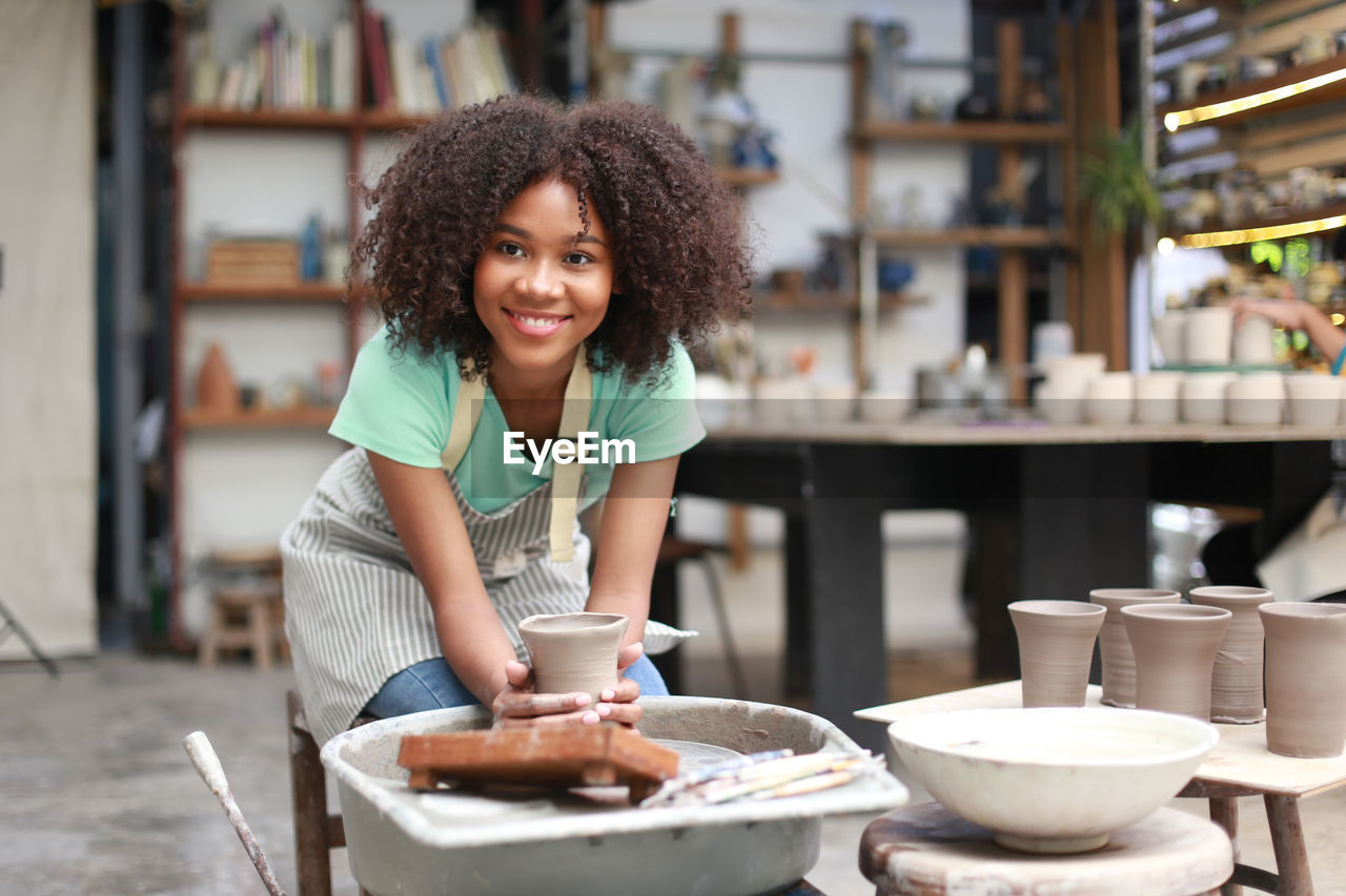 women, adult, smiling, curly hair, one person, happiness, occupation, business, small business, portrait, cheerful, emotion, hairstyle, looking at camera, food and drink, working, young adult, female, indoors, entrepreneur, cafe, person, lifestyles, front view, business finance and industry, food, enjoyment, standing, table, casual clothing, pride, brown hair, creativity, holding, black hair, restaurant, apron, teeth