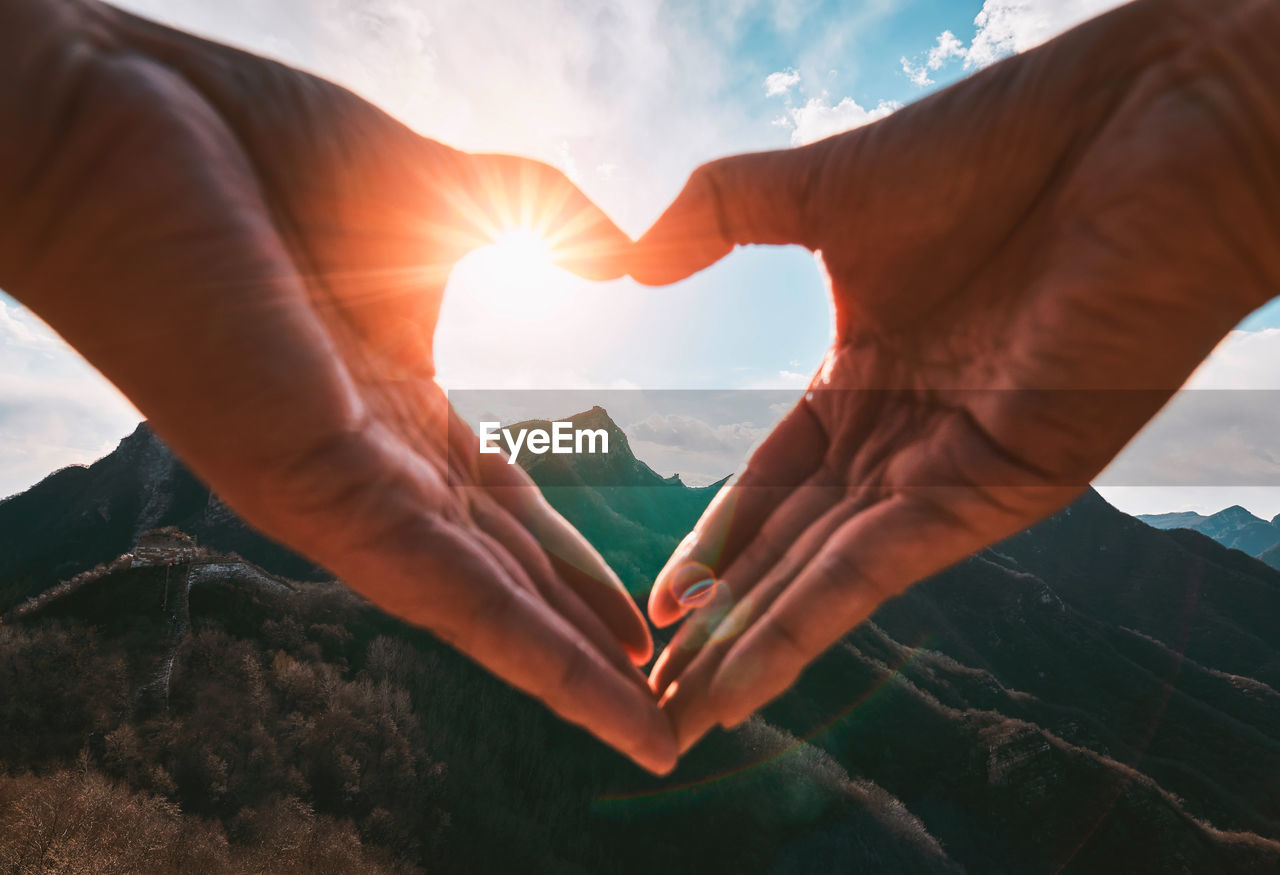 Cropped hands of person making heart shape against mountains and sky