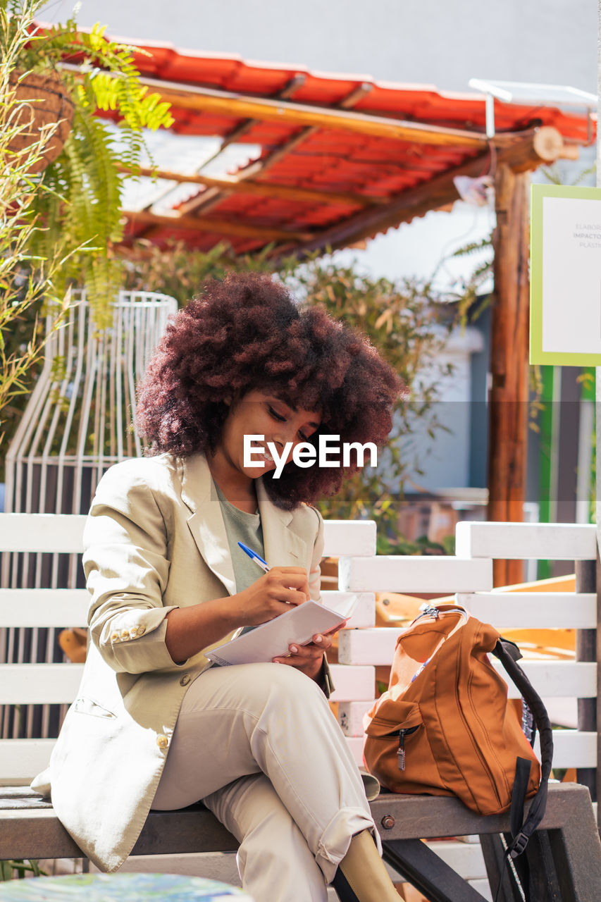 Smiling african american female entrepreneur in suit sitting on bench and writing in planner while working remotely on sunny day in street restaurant