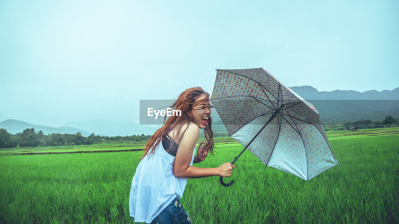 WOMAN WITH UMBRELLA STANDING ON FIELD AGAINST SKY